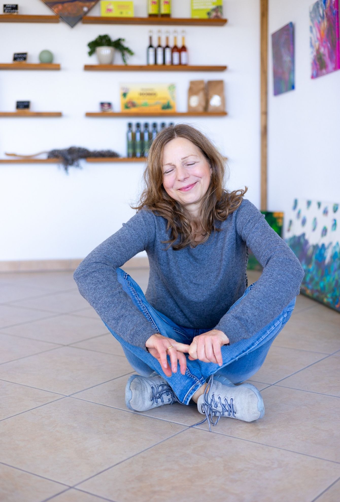 Porträt einer Unternehmerin in lockerer Pose: Sie trägt ein grauen Pullover und Jeans, steht mit den Händen in den Hosentaschen und lächelt freundlich in die Kamera. Im Hintergrund sind Regale mit Ölflaschen und Produkten zu sehen.  Fotografiert in der **Ölmanufaktur Kaltenberg, Region Landsberg am Lech**. Personal Branding und Webseiten Fotografie für die Öl-Manufaktur olio vero in Geltendorf